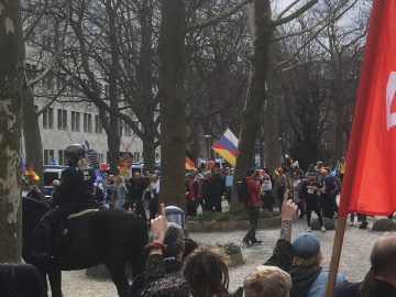Ein Demozug mit alten Menschen. Es sind Deutschlandfahnen und eine diagonal geteilte Fahne mit oben russischer, unten deutscher Trikolore zu sehen. Im Vordergrund eine berittene Polizistin und die Hinterköpfe von Gegendemonstrierenden.